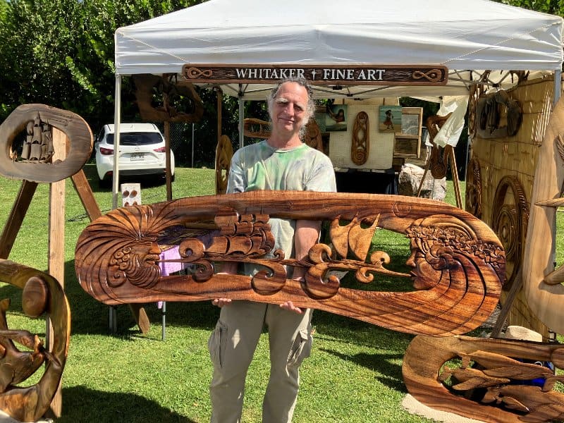 Bruce Whitaker holding a hand-carved koa wave sculpture at his Whitaker Fine Art show tent on Maui